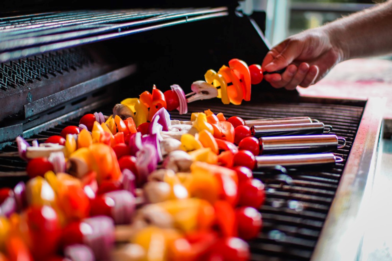 A mans hand laying colorful vegetable kebabs on a gas grill in summer.